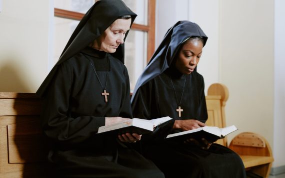 Two ethnically diverse nuns sitting on pew in Catholic church and reading prayer books