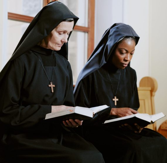 Two ethnically diverse nuns sitting on pew in Catholic church and reading prayer books