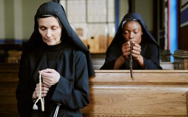 Two ethnically diverse young and mature Catholic nuns sitting on pews in church, praying with rosary in hands